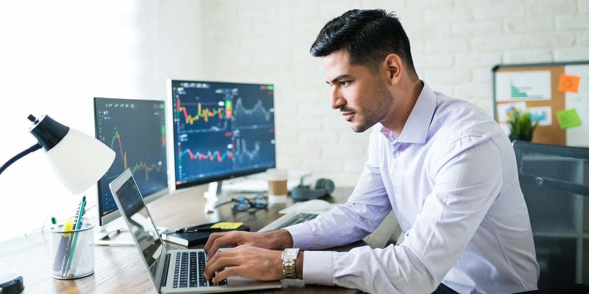 Handsome Latin freelance broker trading through laptop at desk while working from home